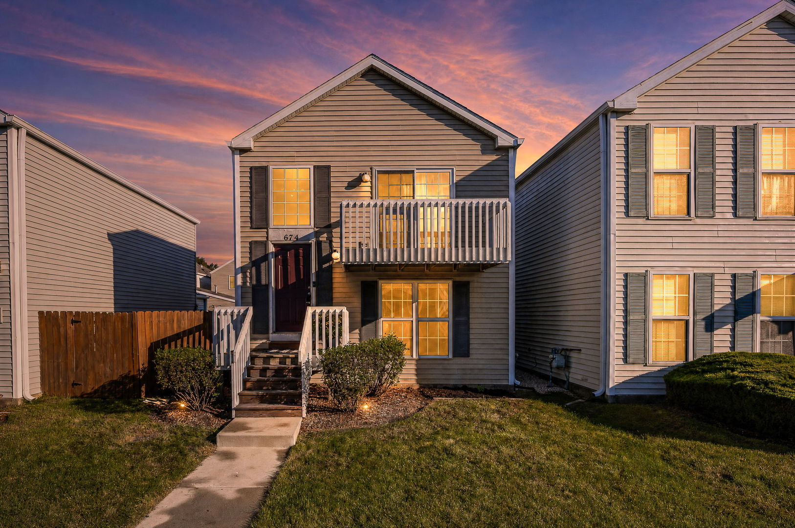 Gray townhouse with a raised entrance and balcony, glowing windows, and a pink dusk sky.