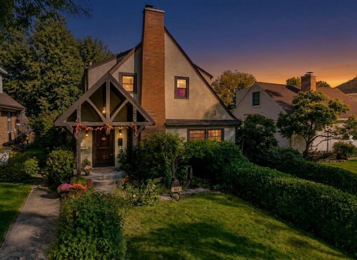 Two-story stucco house with a brick chimney, timber-trimmed porch, and landscaped front garden at dusk.