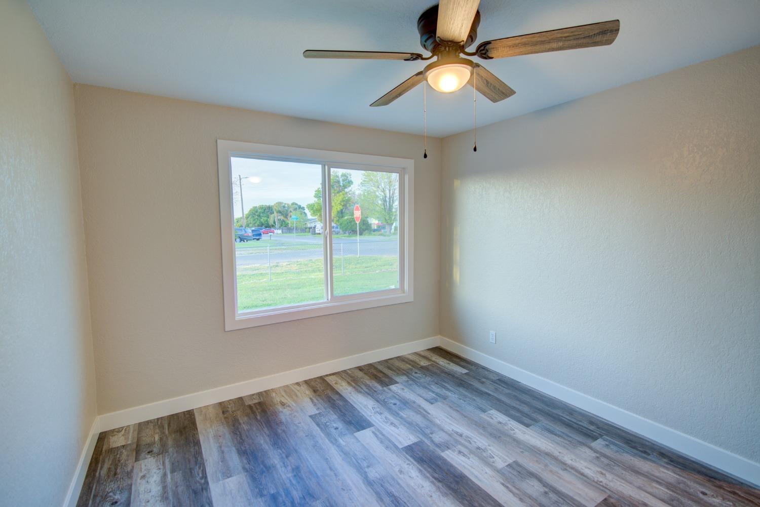Empty bedroom with plank flooring, ceiling fan, and a large window overlooking a grassy street corner.
