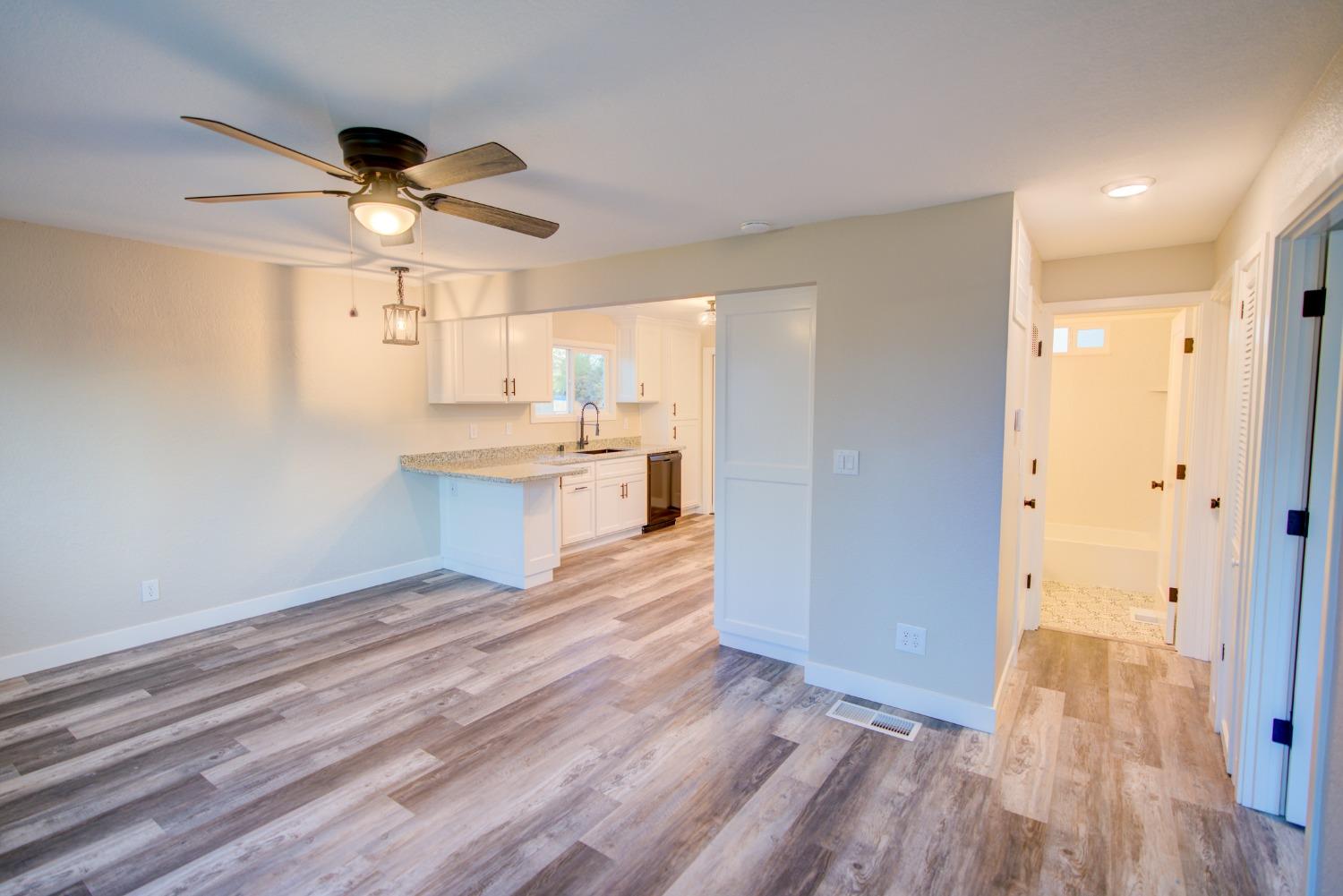 Open-plan room with a white kitchen, granite counters, wood-look flooring, and ceiling fan.