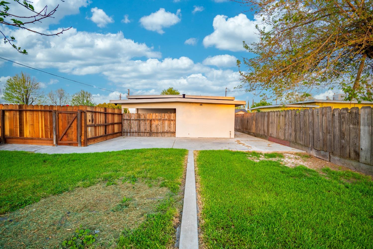 Fenced backyard with green lawn, concrete path, patio, and small outbuilding.