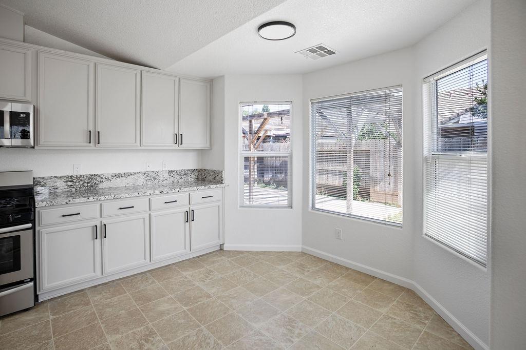 Kitchen with white cabinets, patterned stone countertops, beige tile floor, and three windows with blinds.