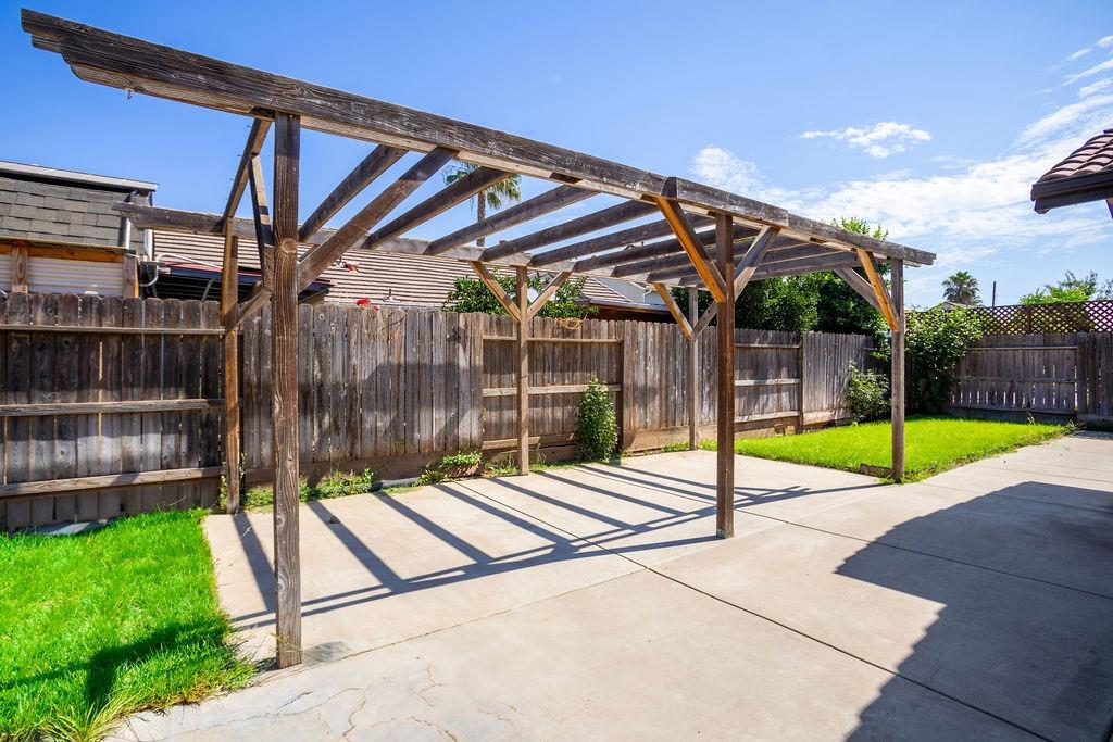 Backyard patio with a large wooden pergola, concrete paving, lawn, and privacy fence.