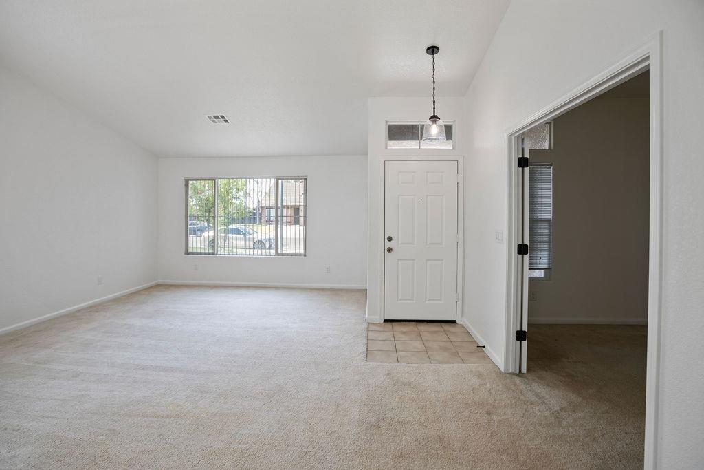 Empty living room with beige carpet, a large barred window, and a tiled entry by the front door.