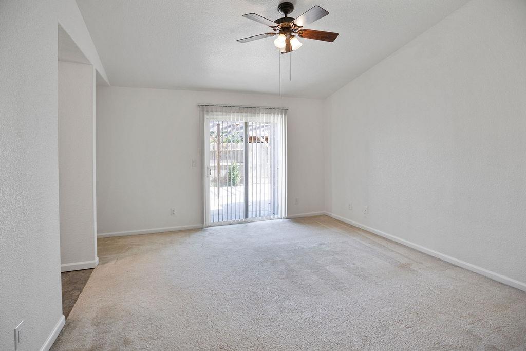 Empty living room with carpet, ceiling fan, and sliding glass door with vertical blinds.