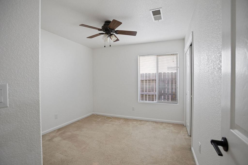 Empty bedroom with beige carpet, ceiling fan, and a window overlooking a fence.