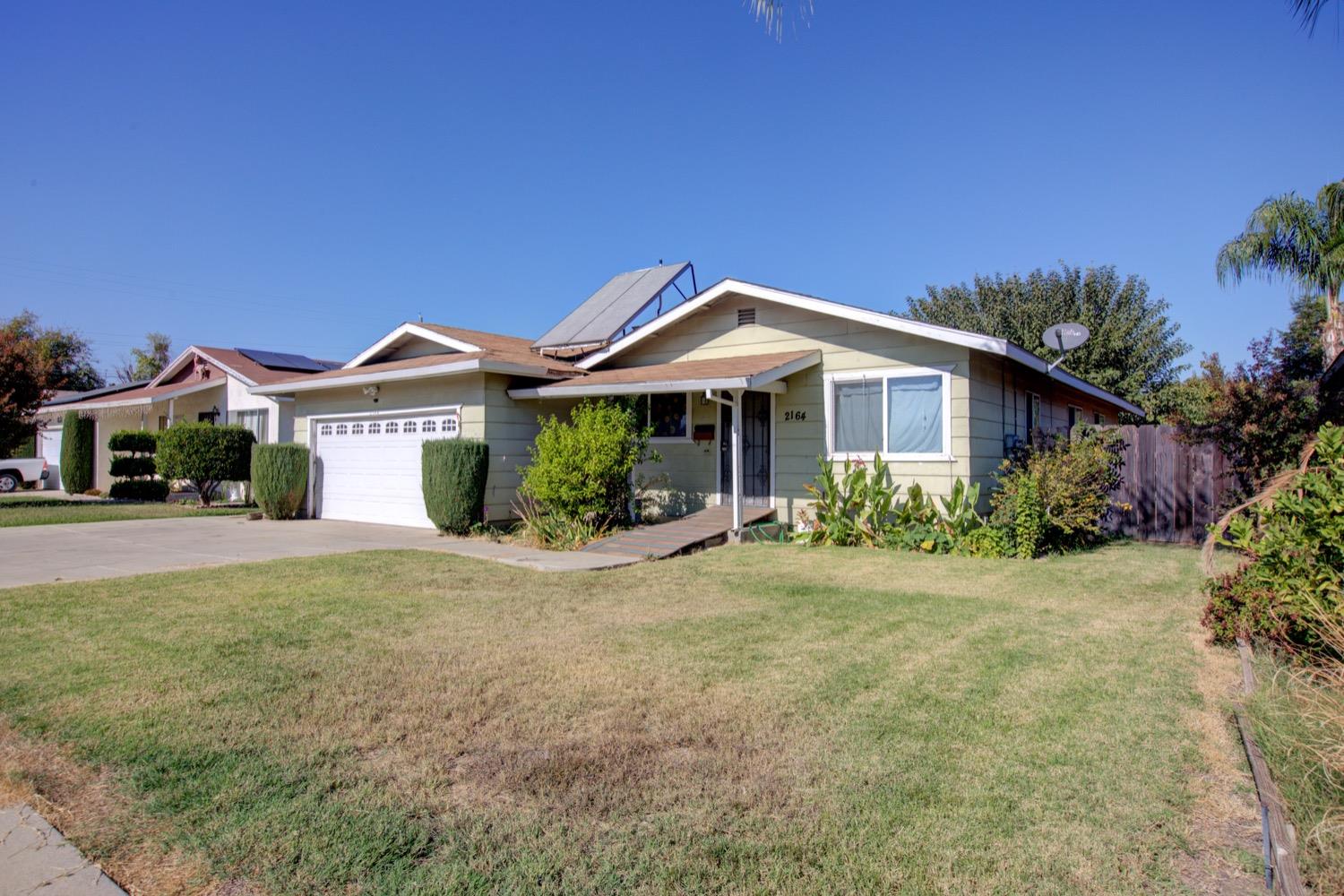 Single-story pale green home with rooftop solar panels, white garage door, and broad front lawn.