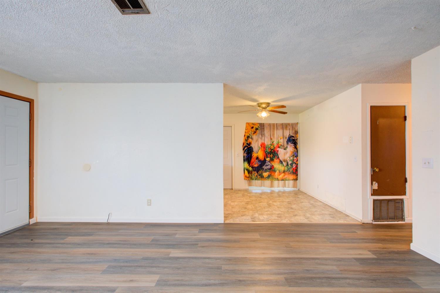 Mostly empty living room with gray plank flooring and an open dining area with a ceiling fan.