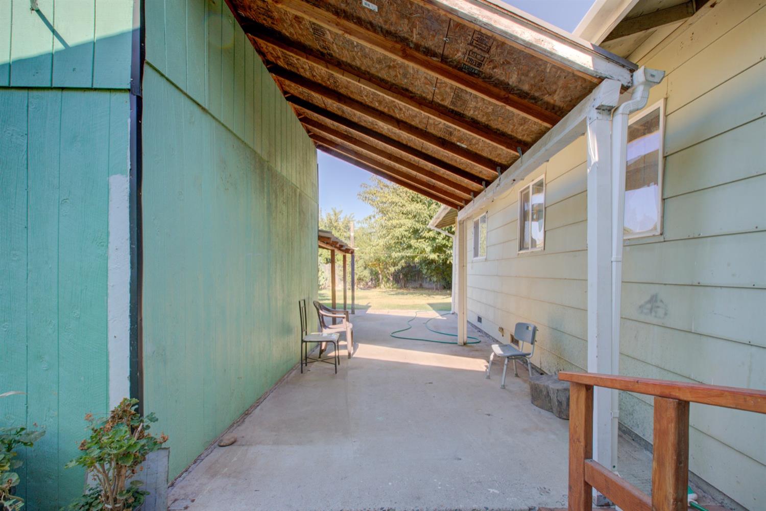 Covered concrete patio with exposed wooden rafters, chairs, and a view of the backyard.