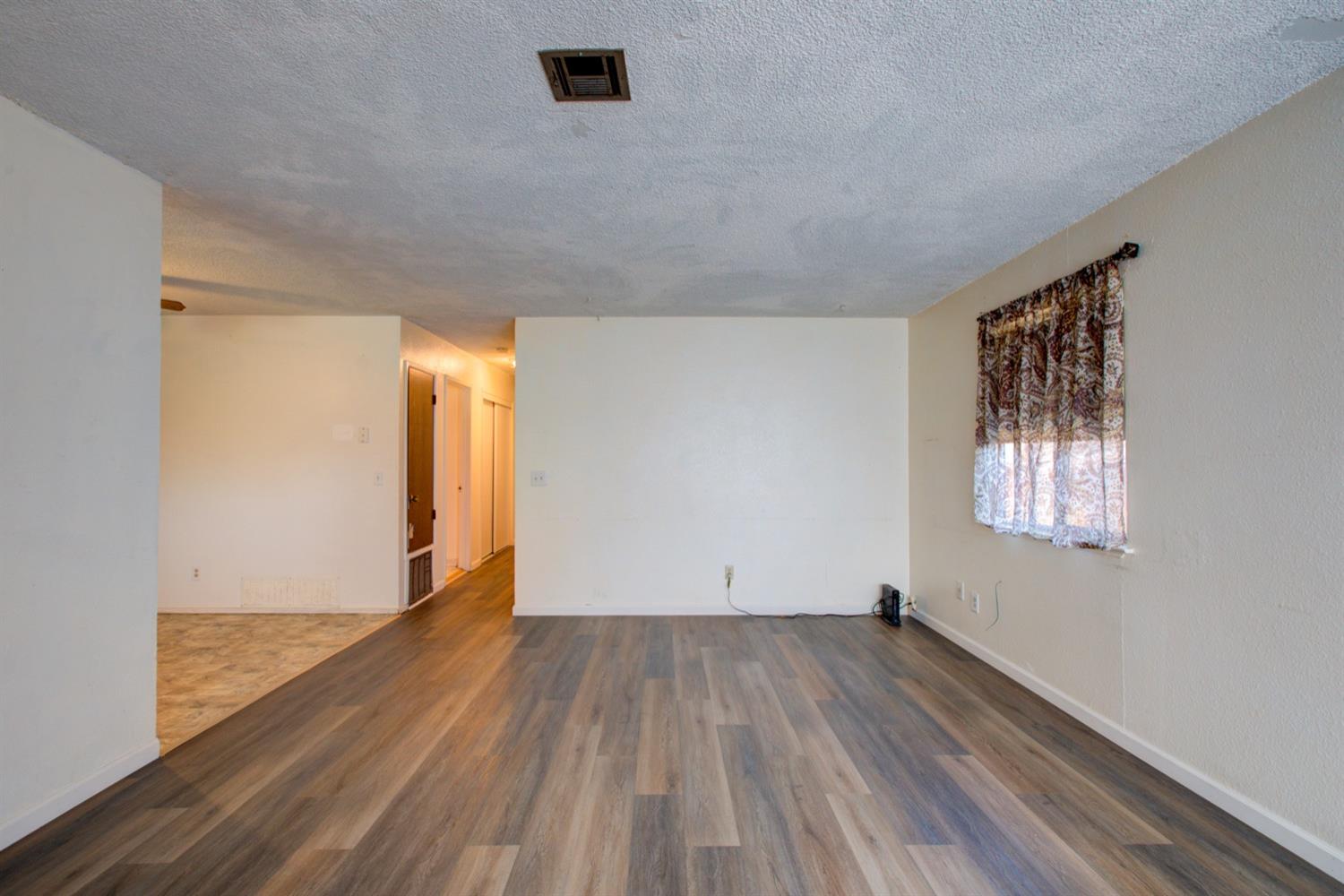 Unfurnished living room with wood-look flooring and a curtained window, opening to a hallway and tiled area.