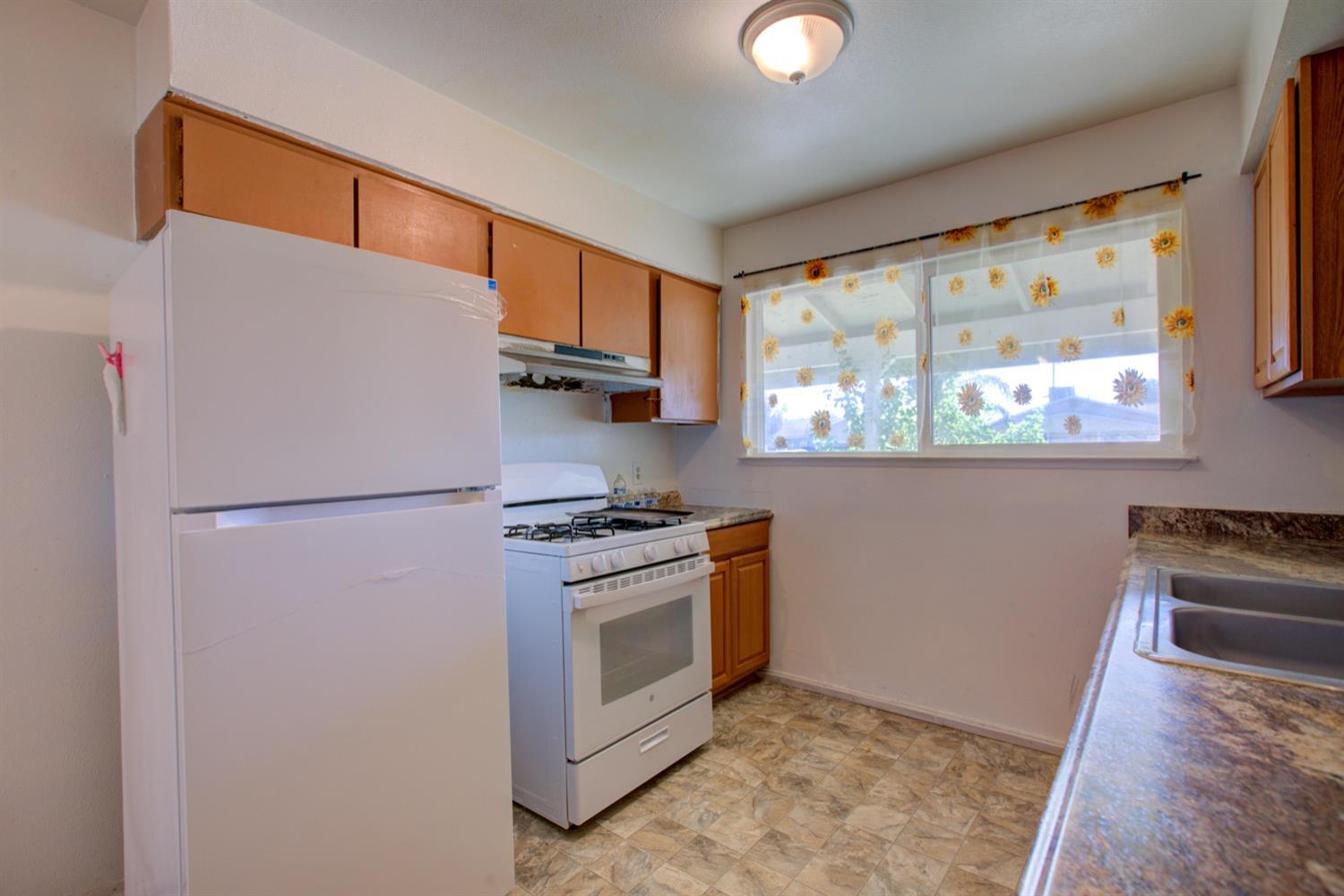 Kitchen with wood cabinets, white refrigerator and gas range, and sunflower curtains at the window.