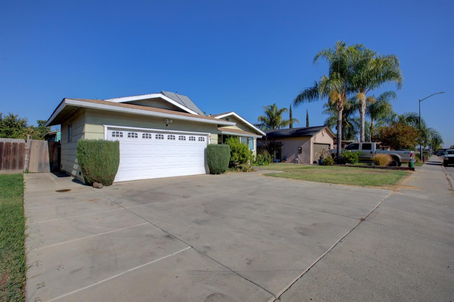 Single-story house with a wide white garage door, broad concrete driveway, and palm trees nearby.
