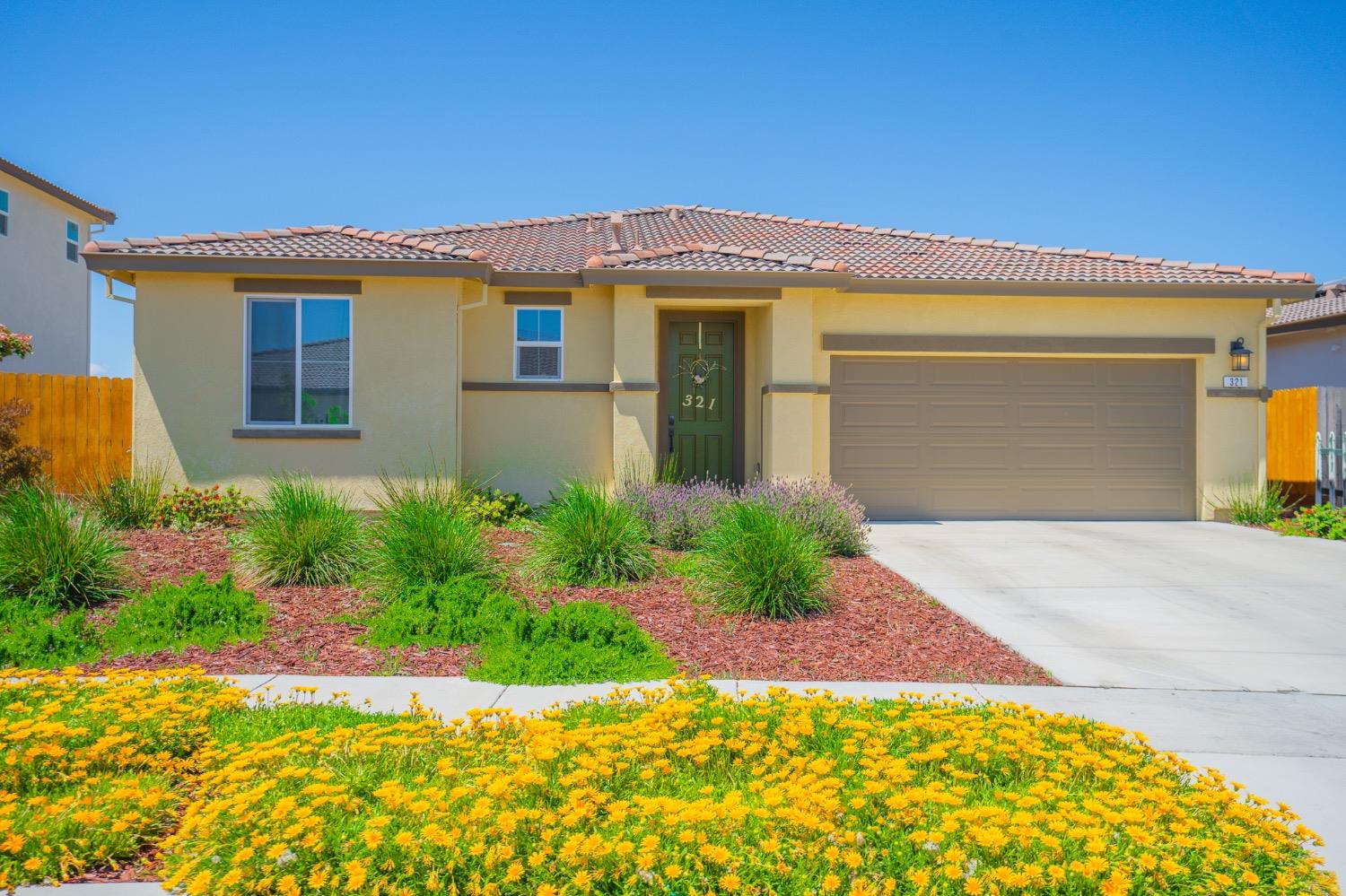Single-story beige home with a tile roof, green front door, two-car garage, and yellow flowers along the driveway.
