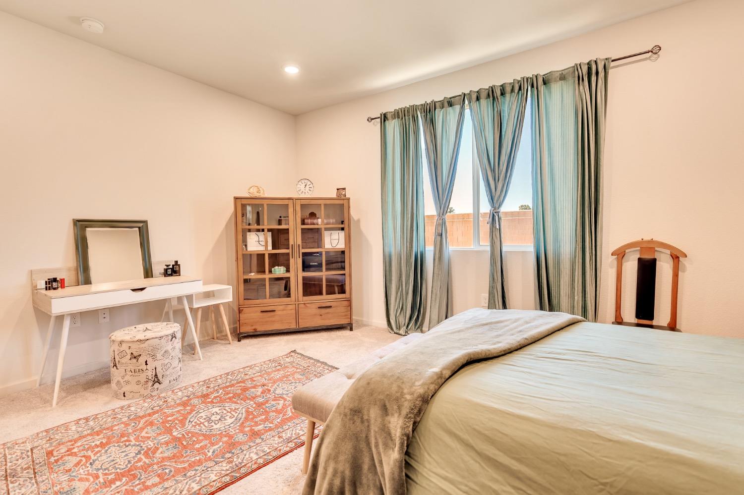 Furnished bedroom with pale green curtains, a white vanity, a wooden display cabinet, and a patterned rug.