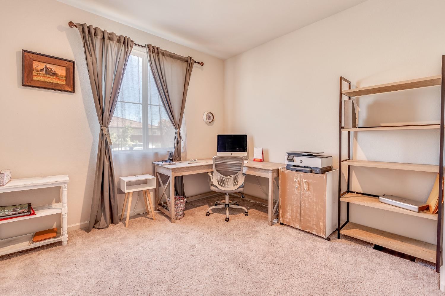 Home office with a corner desk, computer, and open shelving beside a curtained window.