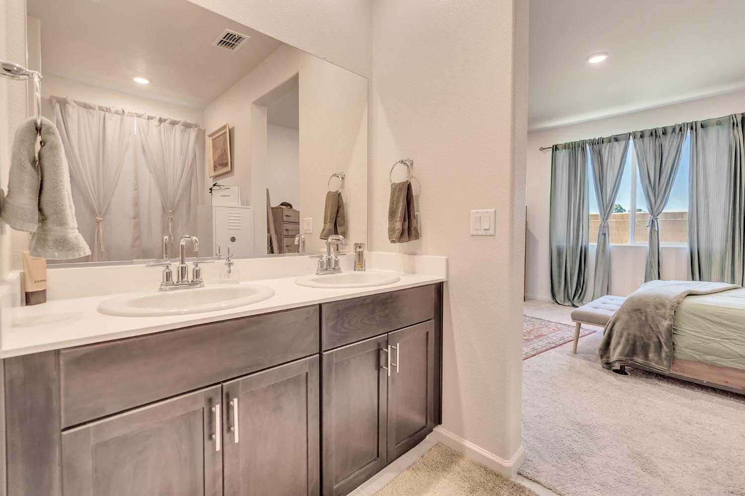 Bathroom with a double-sink vanity and dark cabinets, opening onto a bedroom with curtained windows.
