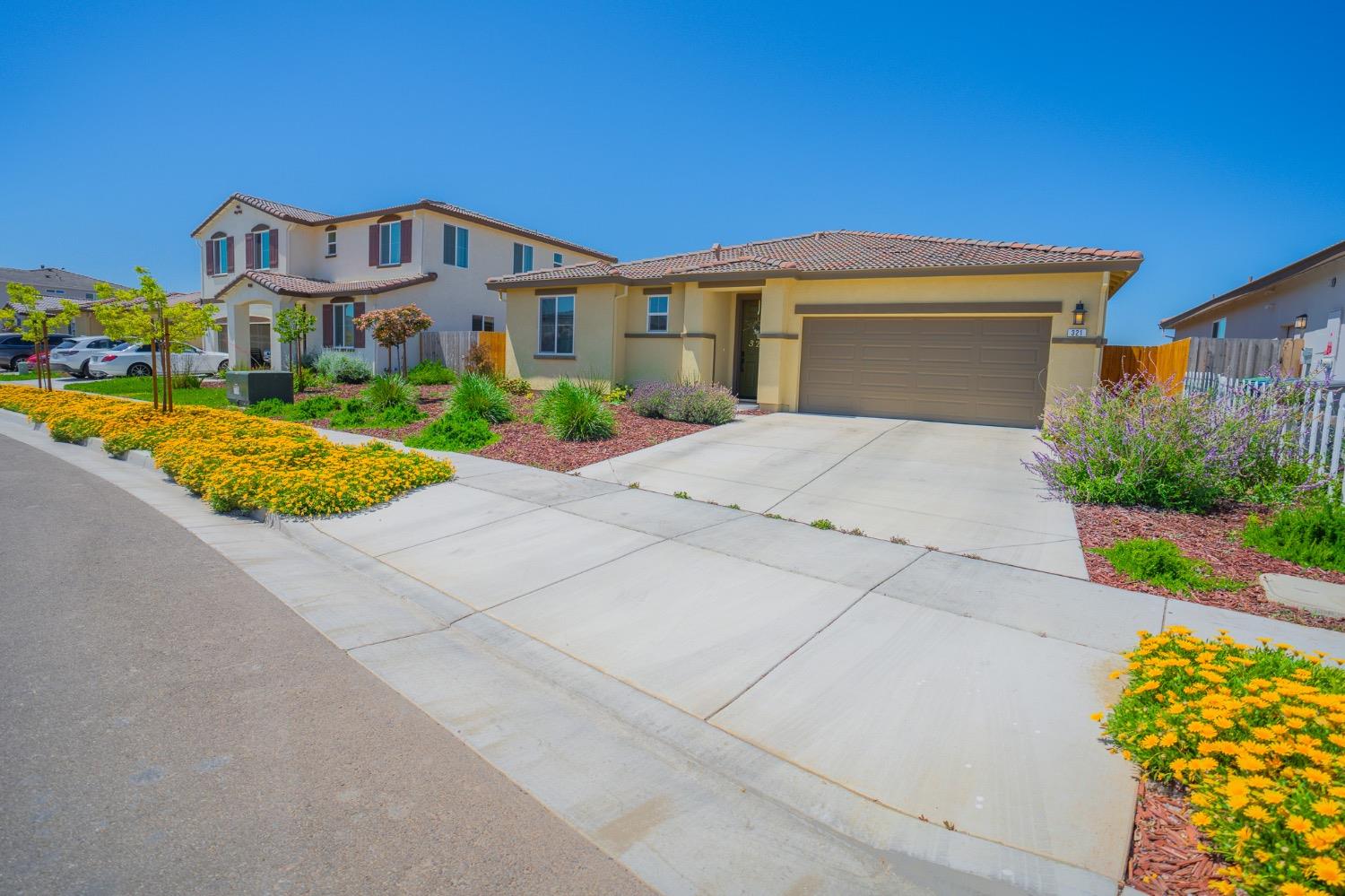 Beige stucco home with an attached two-car garage, broad driveway, and colorful flowering landscaping.