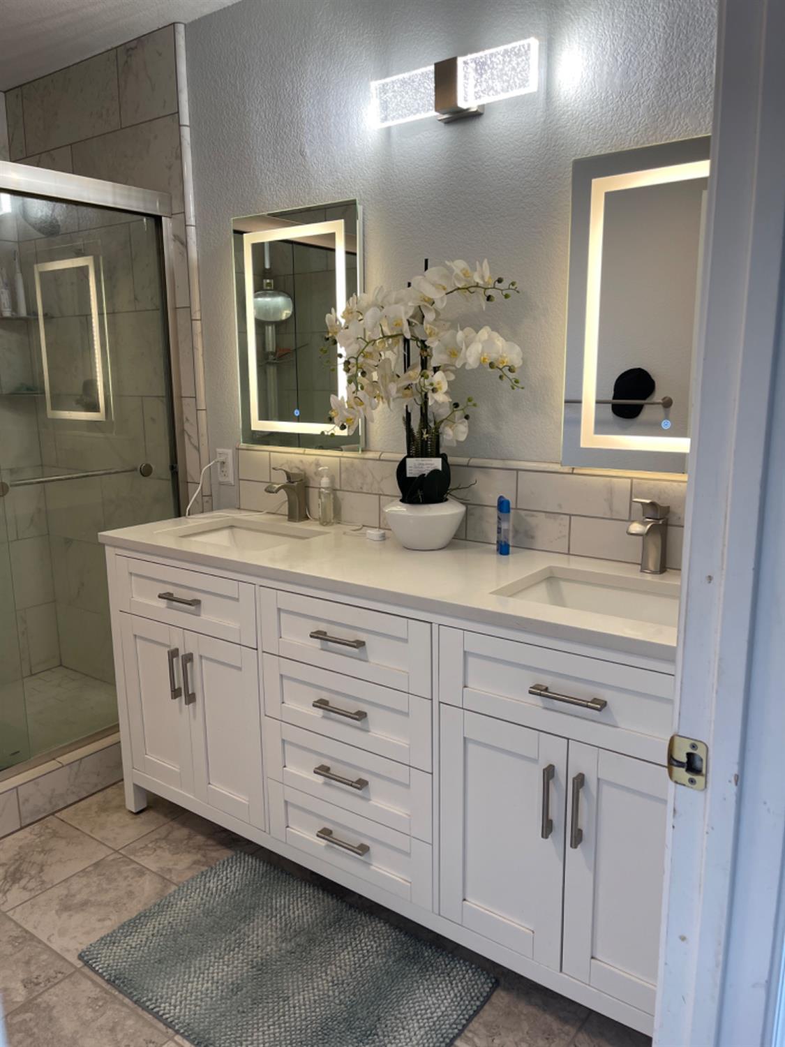 Bathroom with a white double vanity, illuminated mirrors, and a glass-enclosed tile shower.
