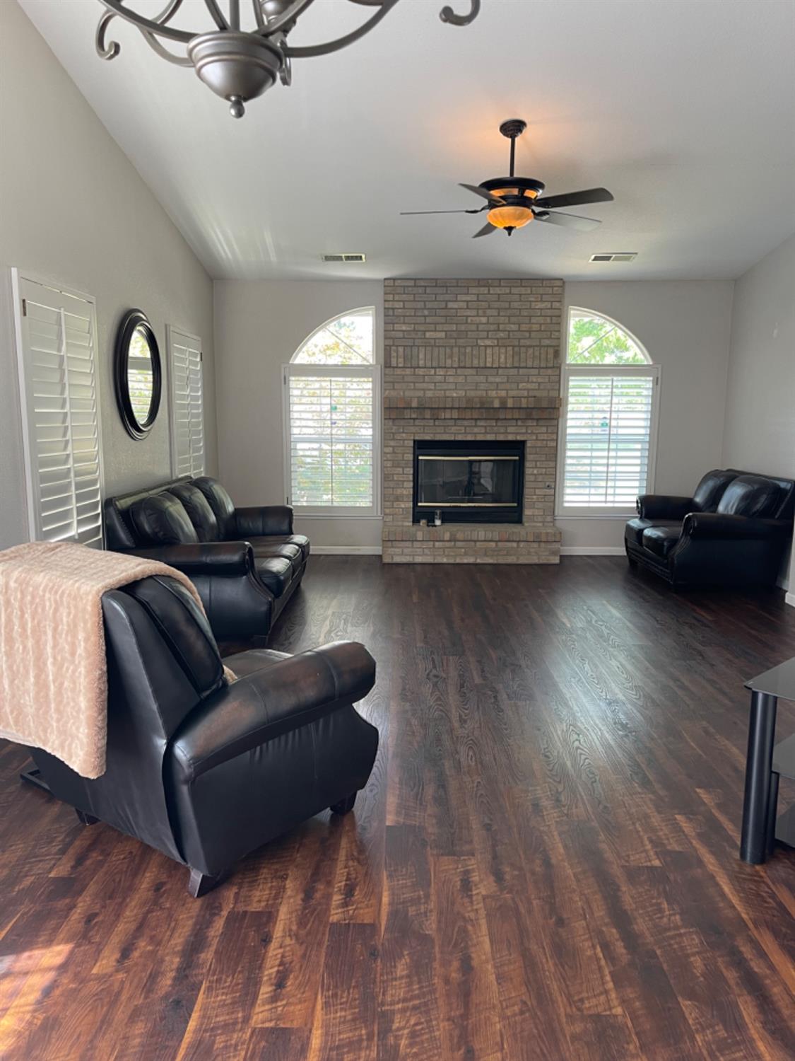 Living room with a brick fireplace, arched shuttered windows, dark wood floors, and black leather seating.