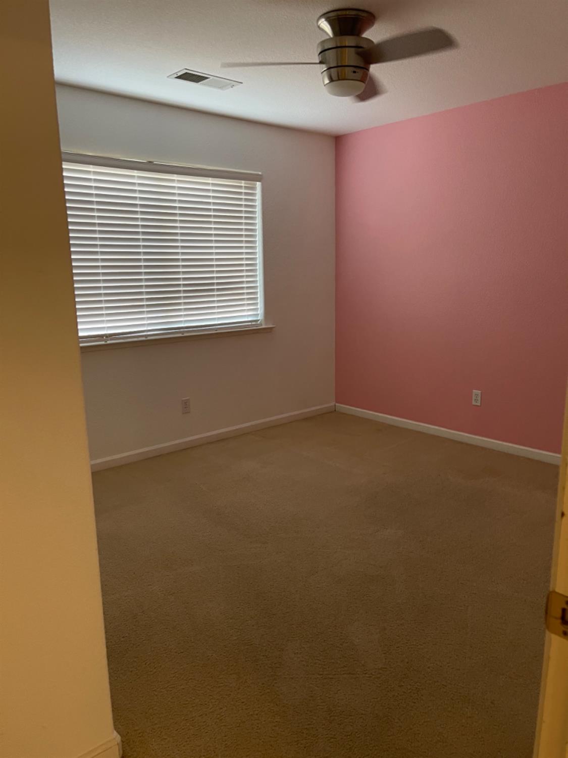 Empty bedroom with beige carpet, pink accent wall, closed blinds, and a ceiling fan.