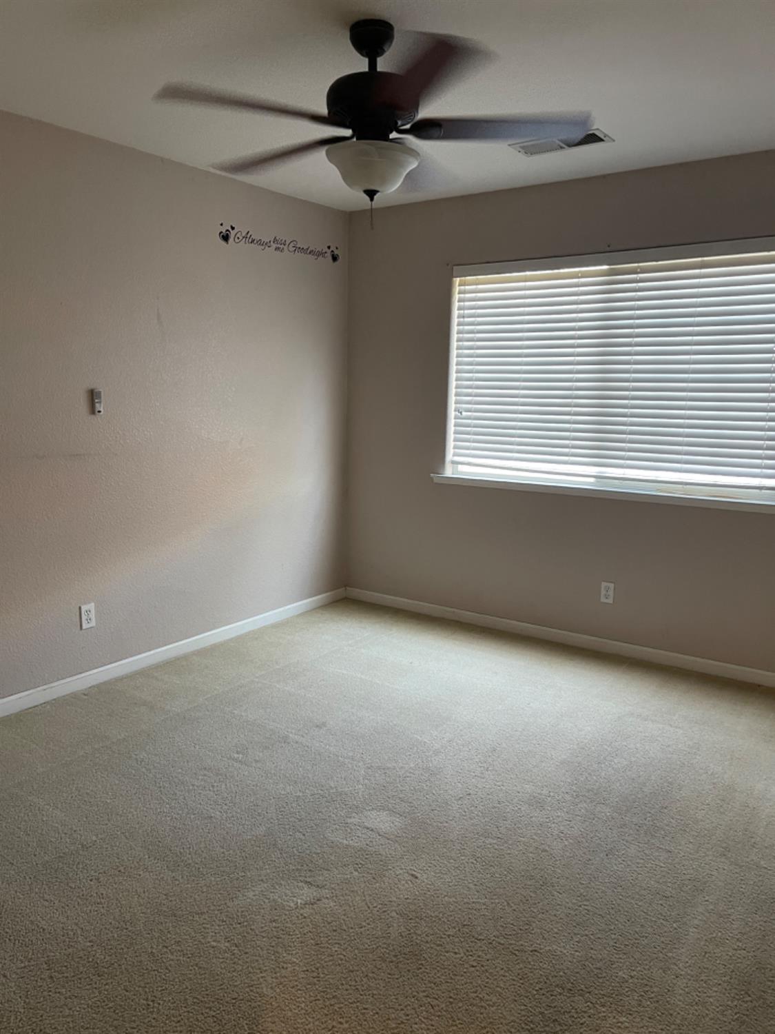 Empty carpeted bedroom with a ceiling fan and a wide window with closed blinds.