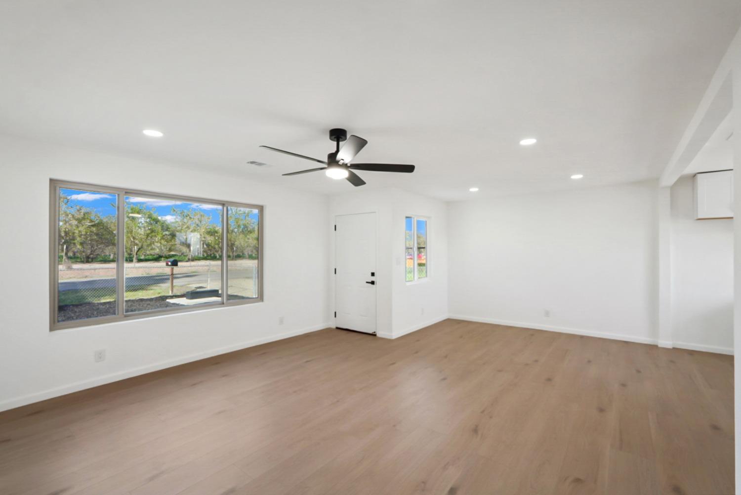 Vacant living room with light wood flooring, a large window, and a black ceiling fan.
