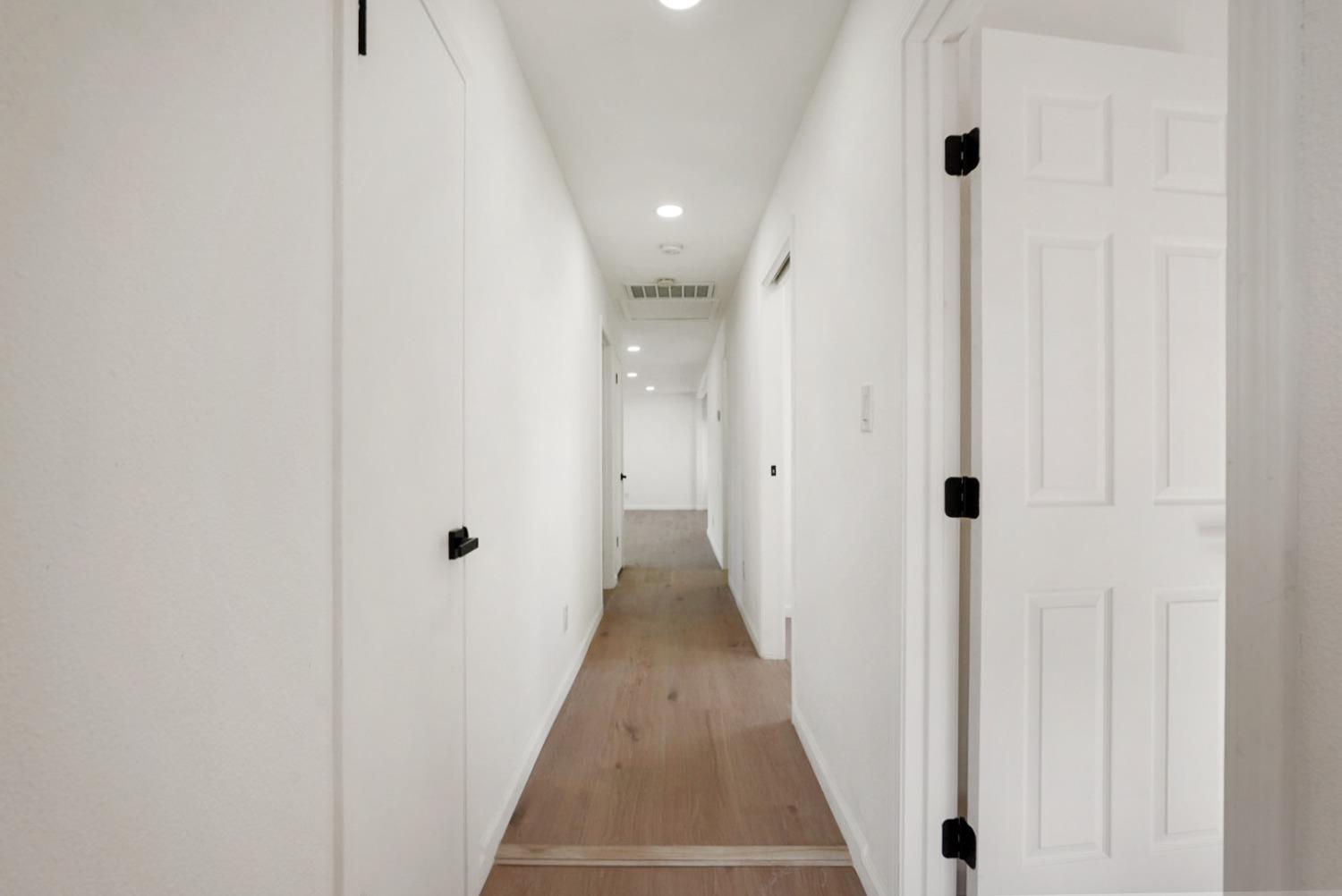White hallway with light wood flooring, recessed lights, and several doors.