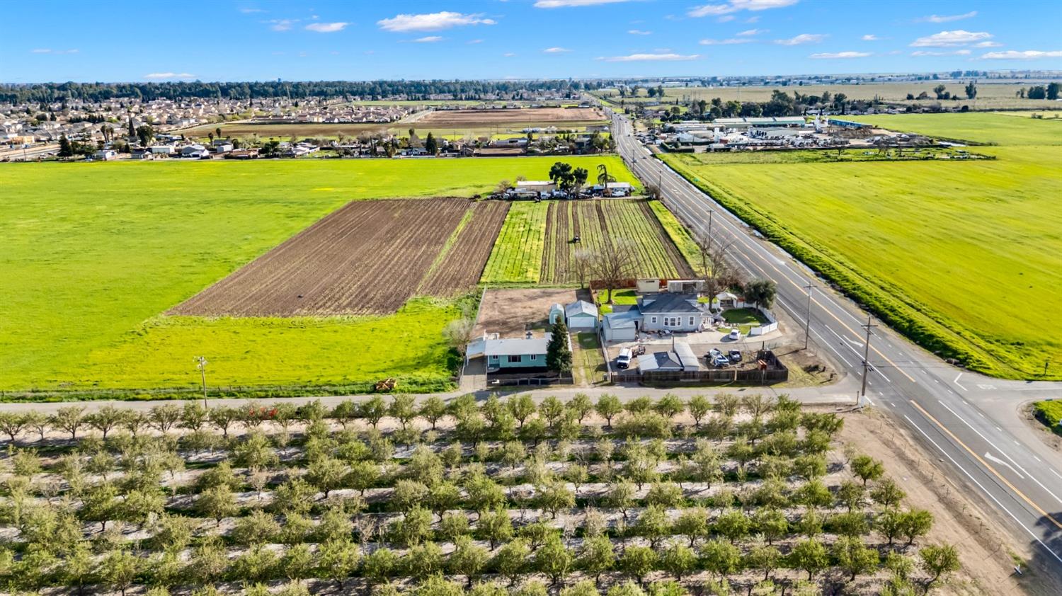 Aerial view of a house and outbuildings amid cultivated fields and orchard rows beside a major road.