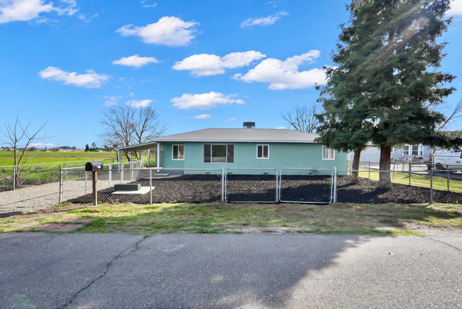 Single-story teal house behind a chain-link fence, with a large evergreen beside it.