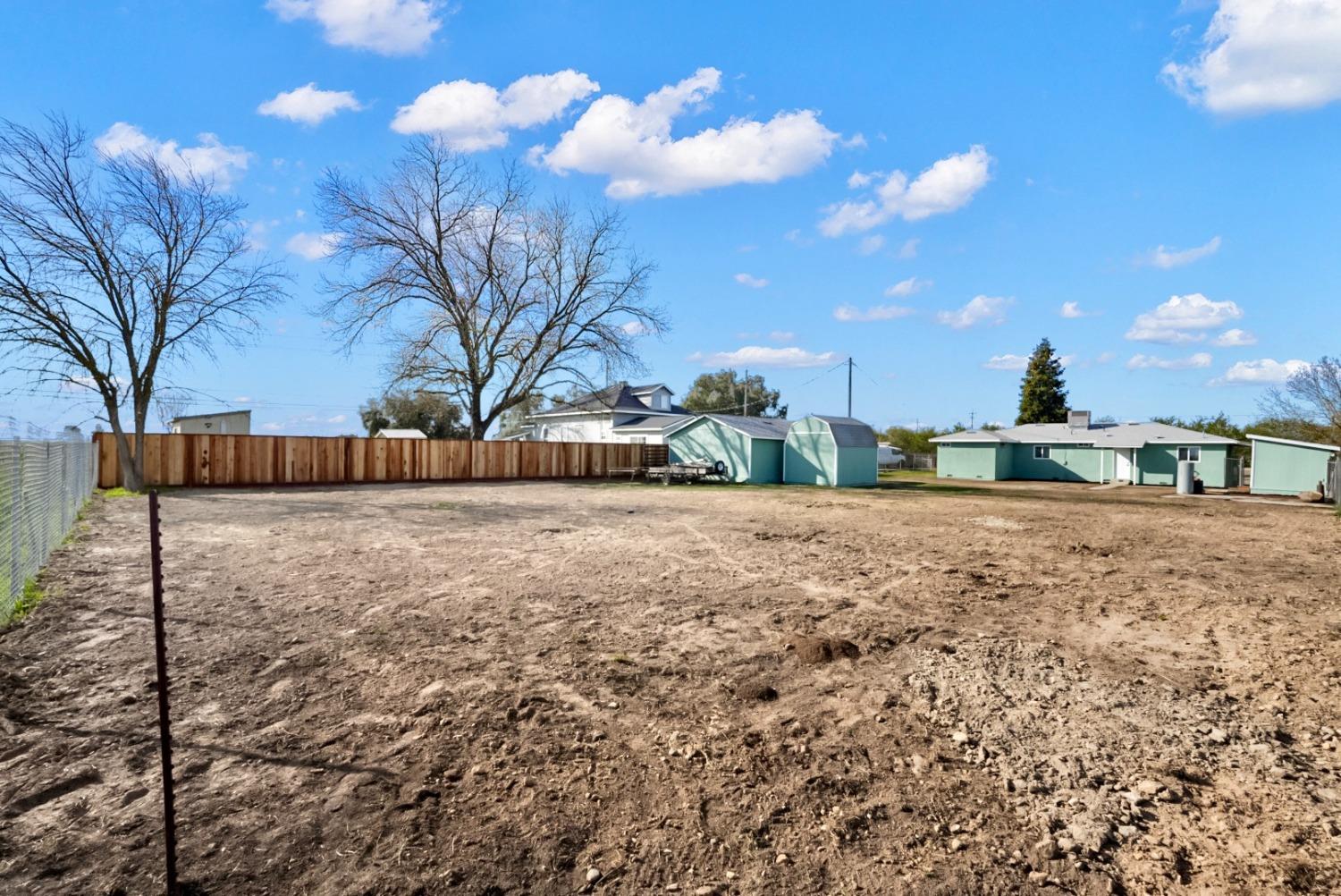 Cleared dirt yard bordered by fencing, with several sheds and a house beyond it.