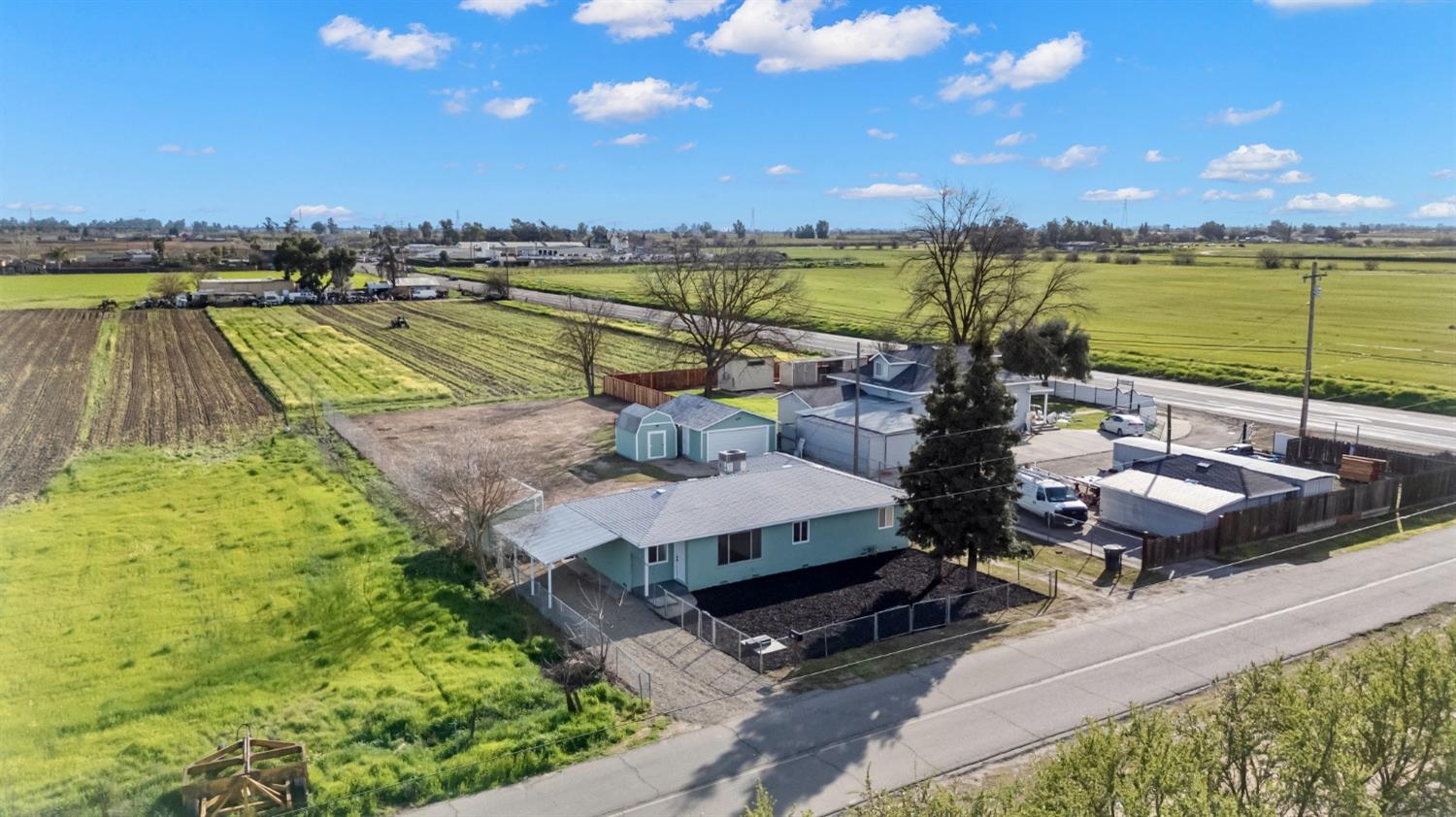 Aerial view of a blue single-story home and outbuildings surrounded by green farmland.