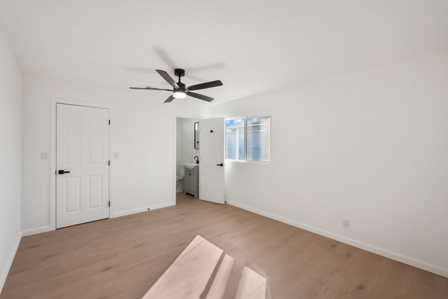 Vacant bedroom with light wood floors, a black ceiling fan, and an open doorway to the bathroom.