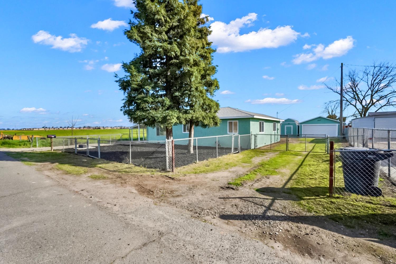 Teal single-story house behind a large evergreen tree and chain-link fence, with garages and farmland nearby.