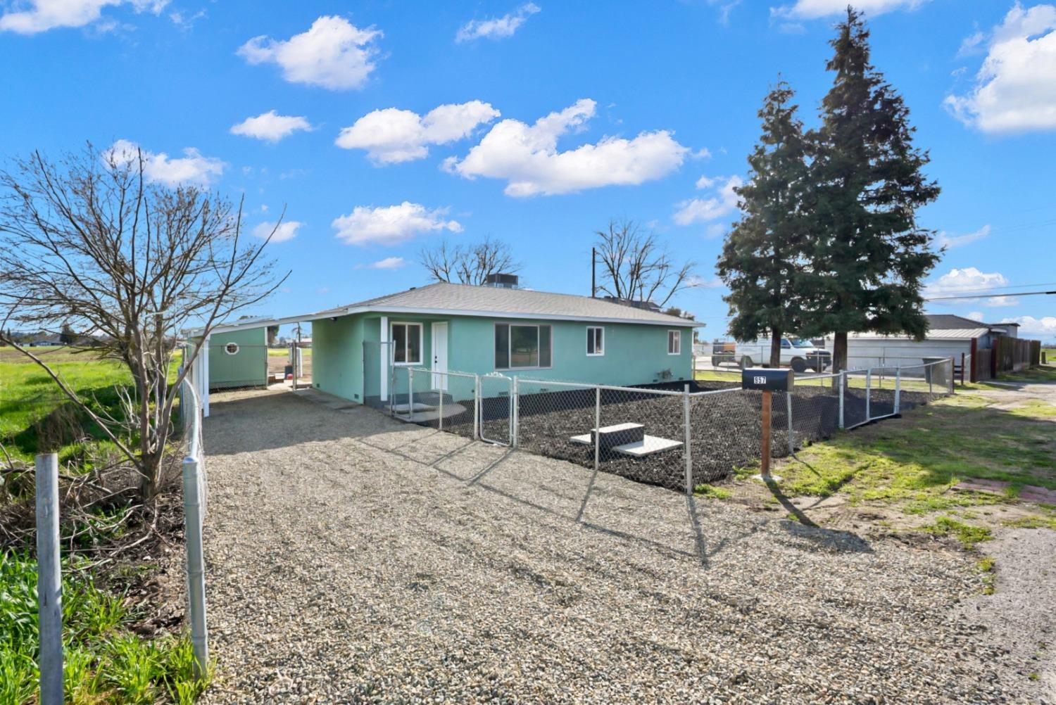 Turquoise single-story house behind a chain-link fence, with a gravel driveway and tall evergreen.