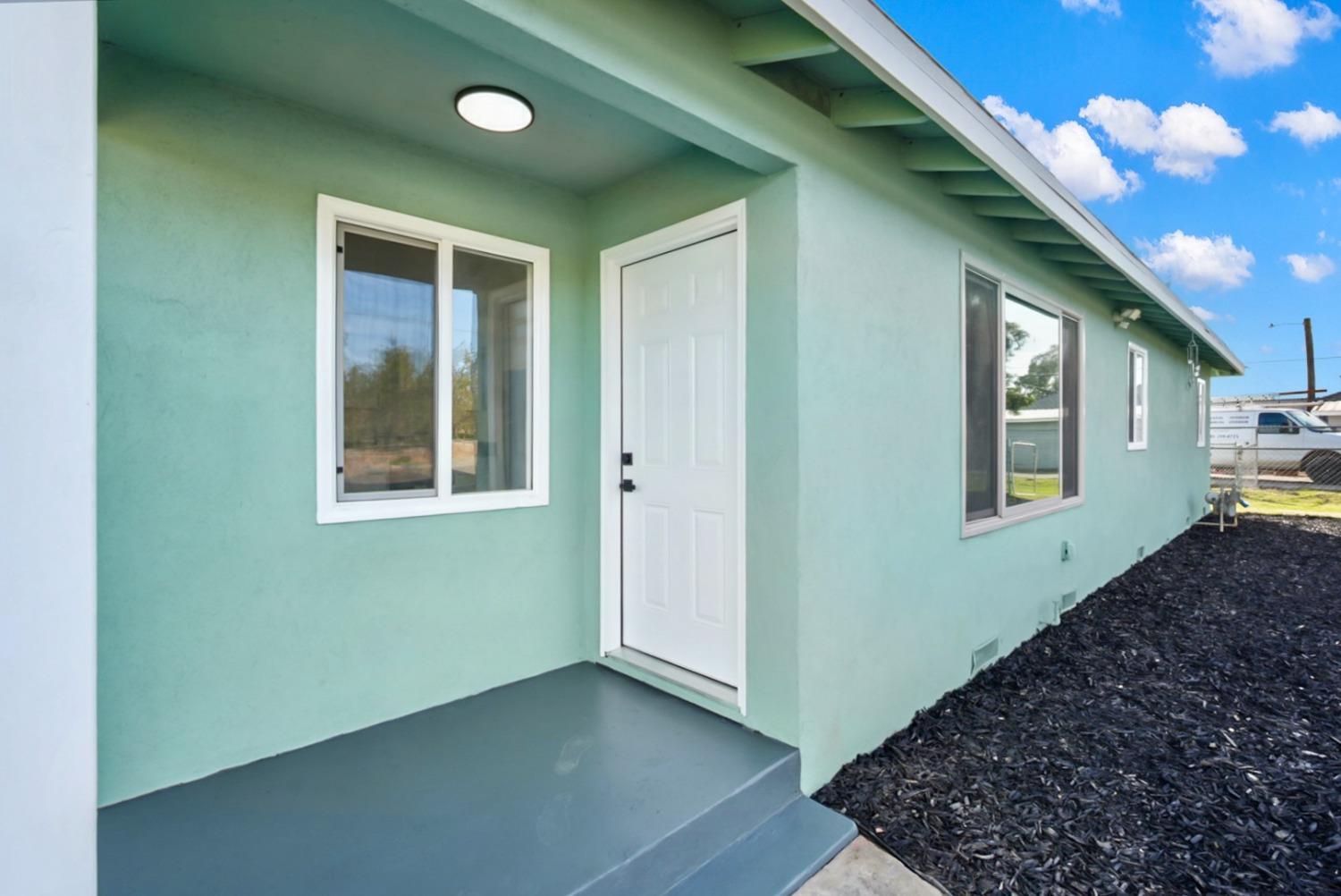 Mint-green house entry with a covered porch, white door, gray steps, and dark mulch landscaping.