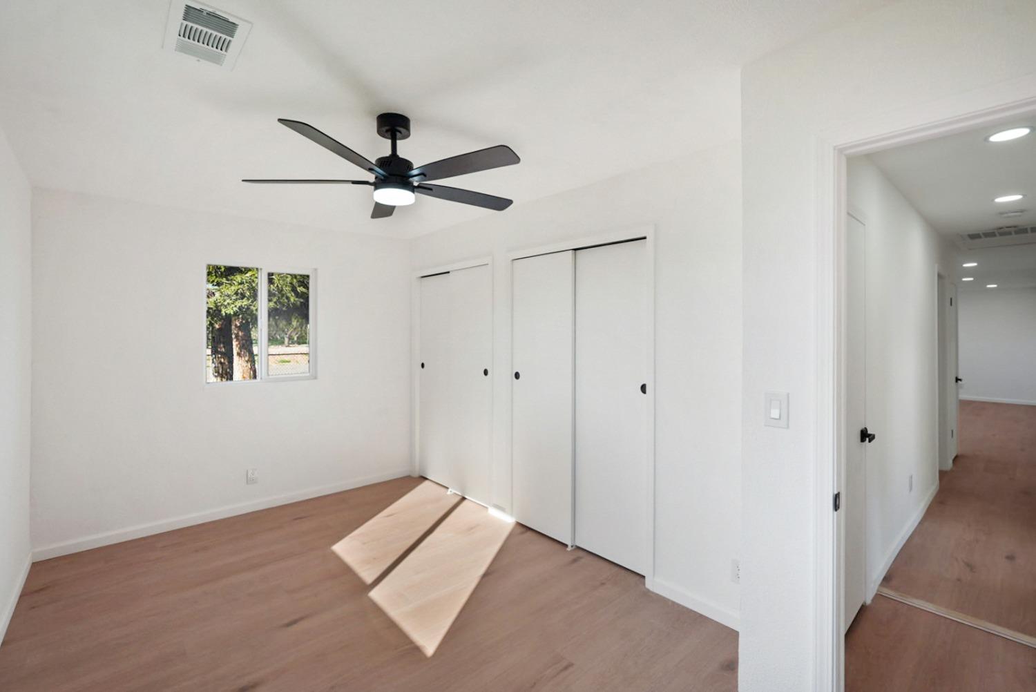 Empty bedroom with sliding closet doors, a black ceiling fan, and sunlight across the wood floor.