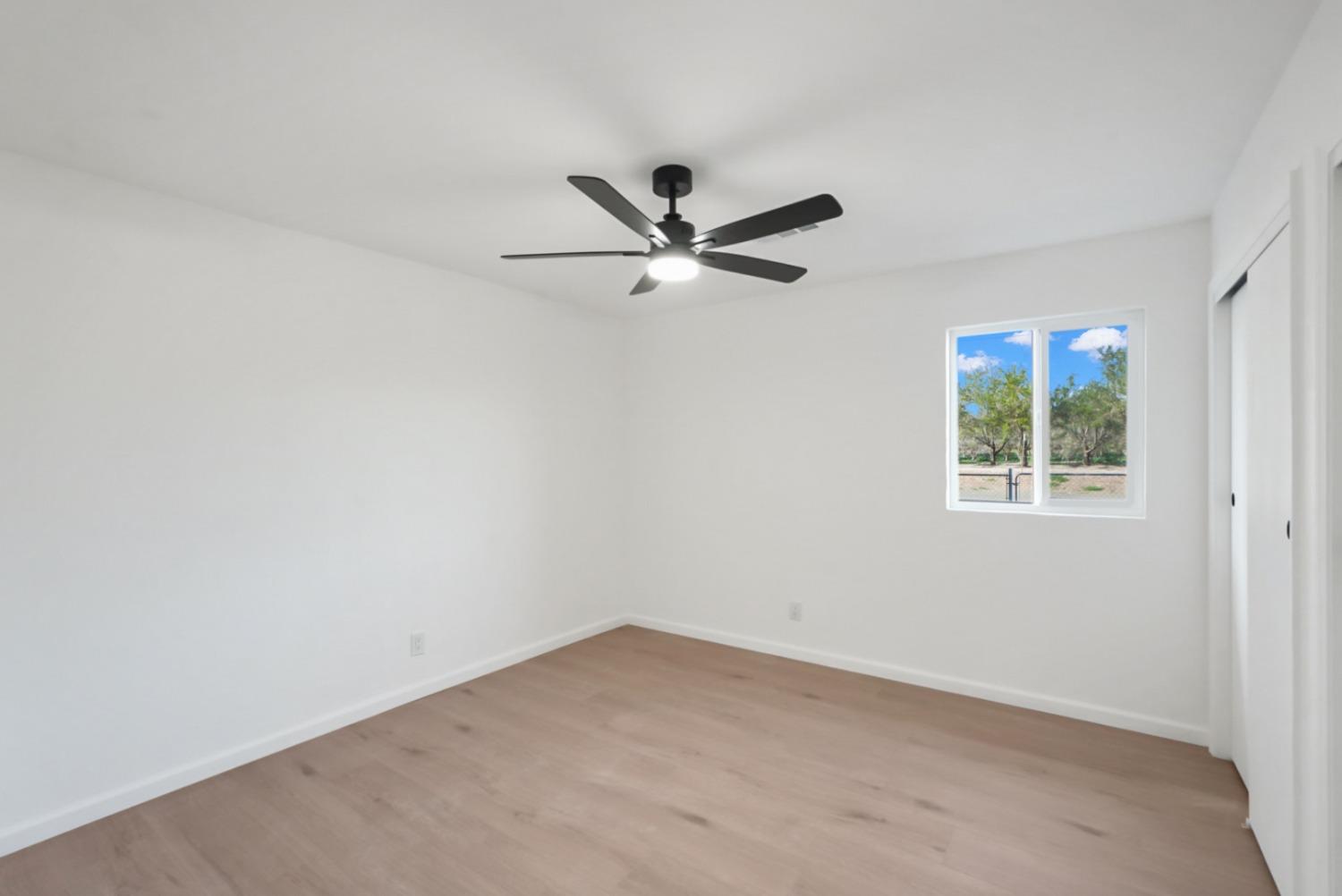 Empty bedroom with light wood flooring, a black ceiling fan, and a window overlooking trees.