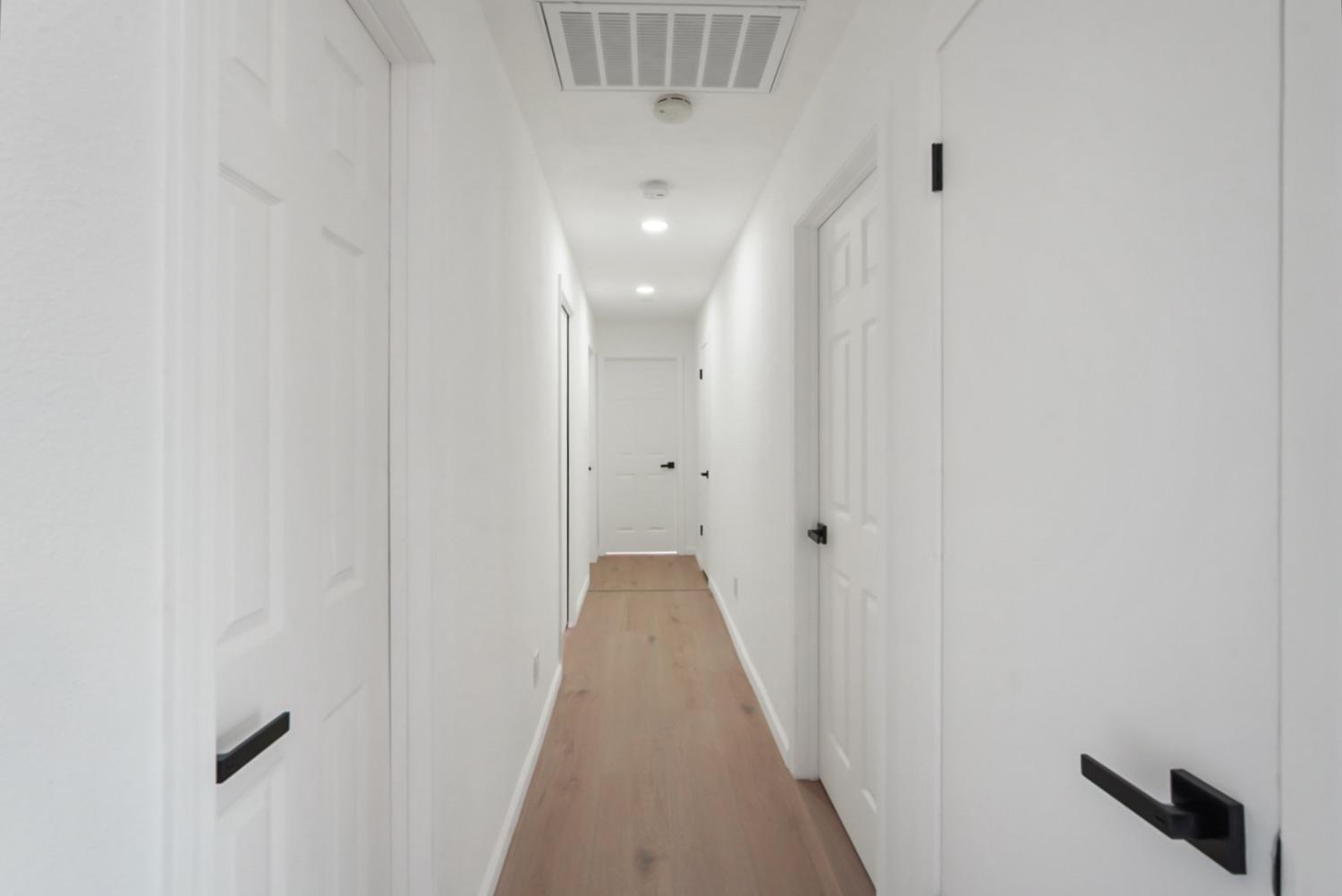 White hallway with light wood flooring, paneled doors, and recessed ceiling lights.