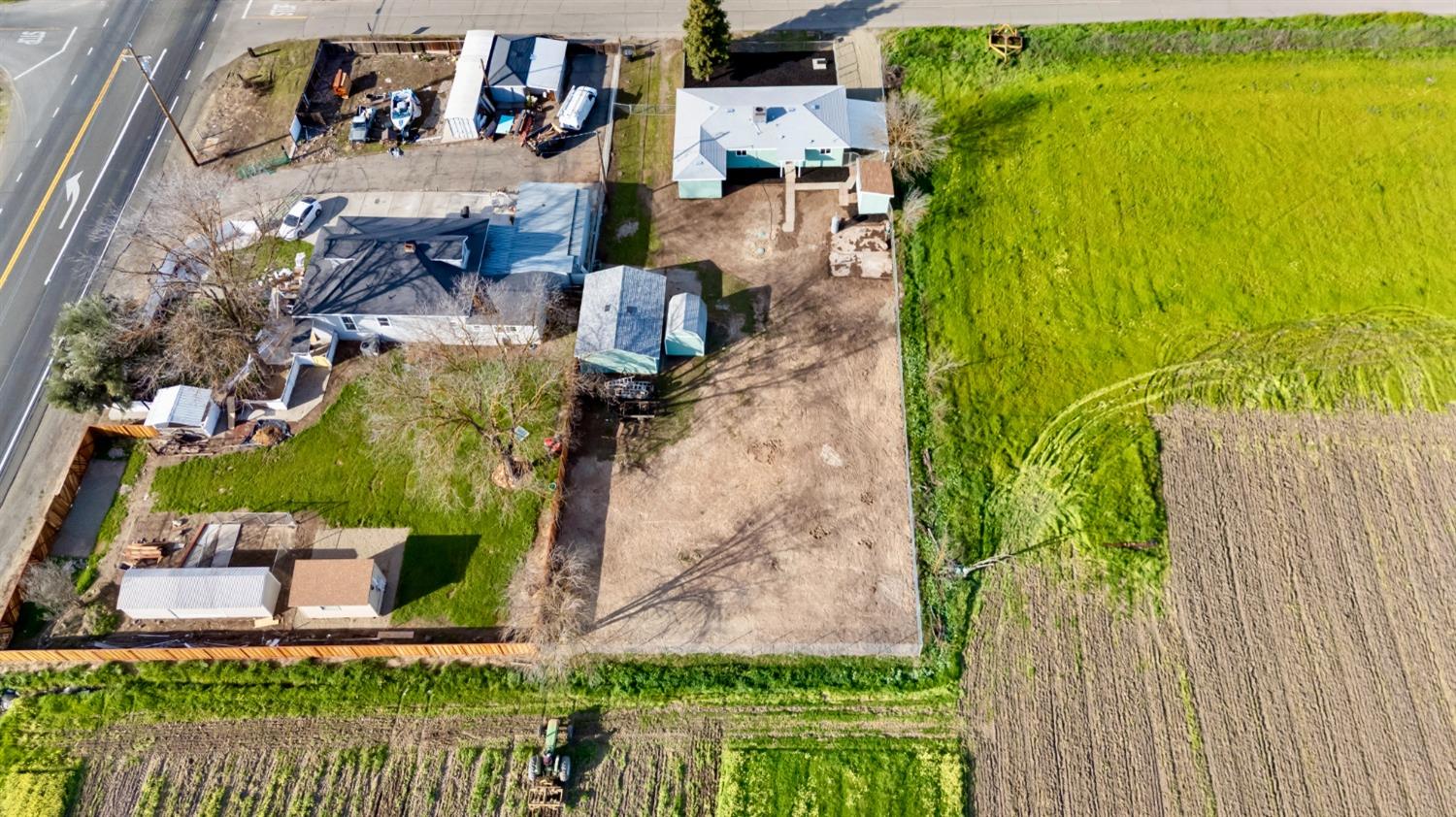 Aerial view of homes and outbuildings beside a cleared dirt lot and green fields.