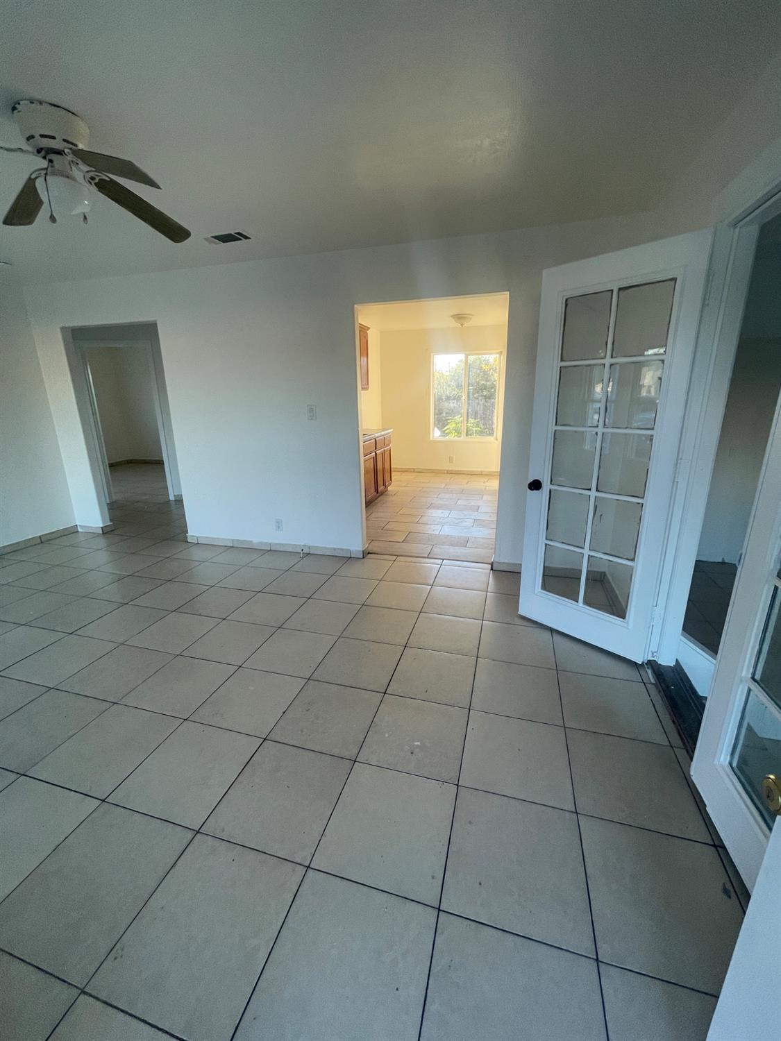Vacant tiled room with a ceiling fan, open French doors, and a bright kitchen beyond.