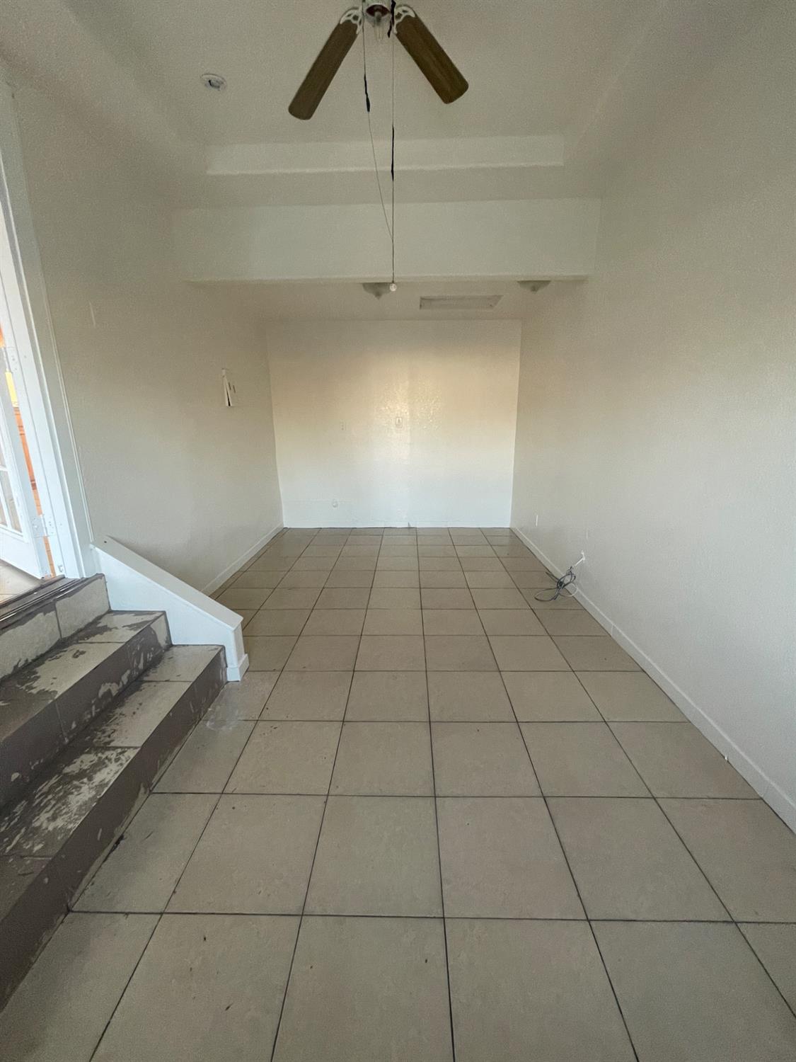 Empty tiled room with a ceiling fan and worn steps beside a bright doorway.