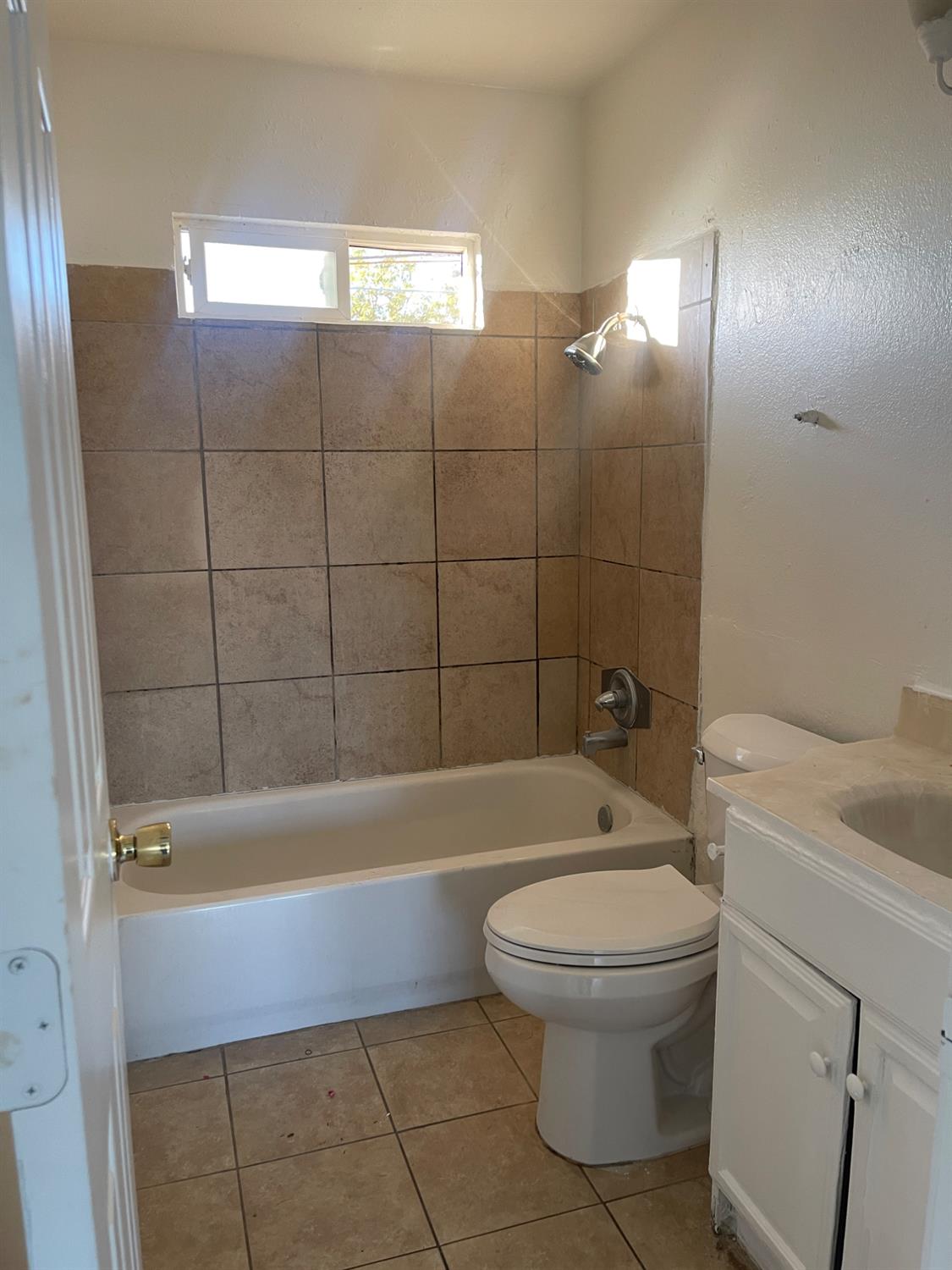 Bathroom with a tan tiled tub and shower, toilet, white vanity, and high window.