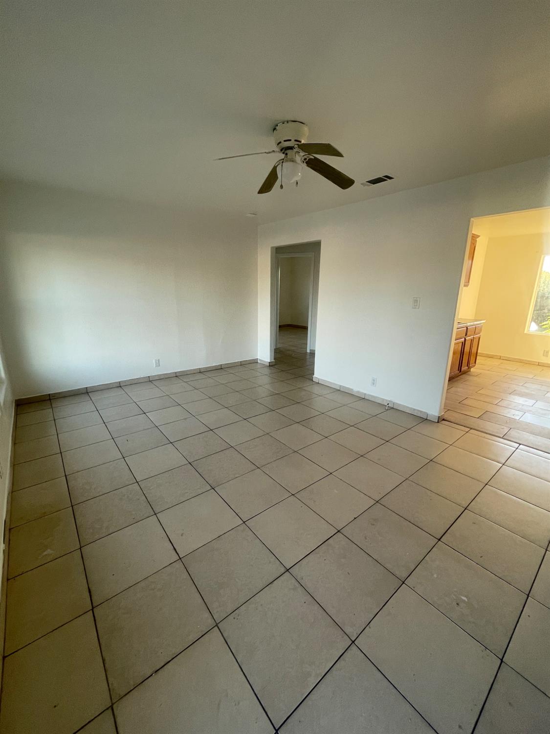 Empty tiled living area with a ceiling fan and an opening into the kitchen.