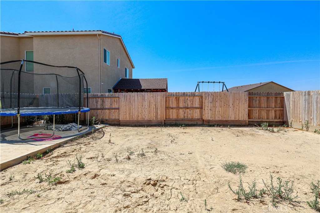 Backyard with dry, bare ground, a wooden fence, and a trampoline on a concrete patio.