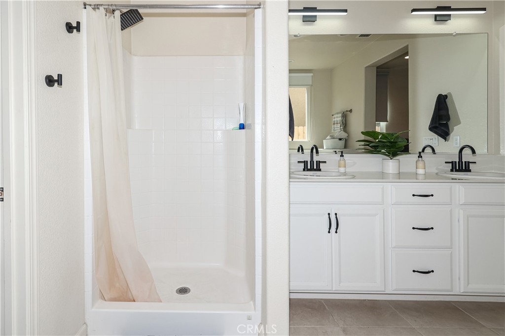 Bathroom with a white tiled shower beside a double vanity with black faucets and a large mirror.