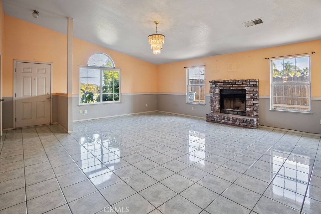 Empty living room with glossy tile floors, a brick fireplace, large windows, and a chandelier.