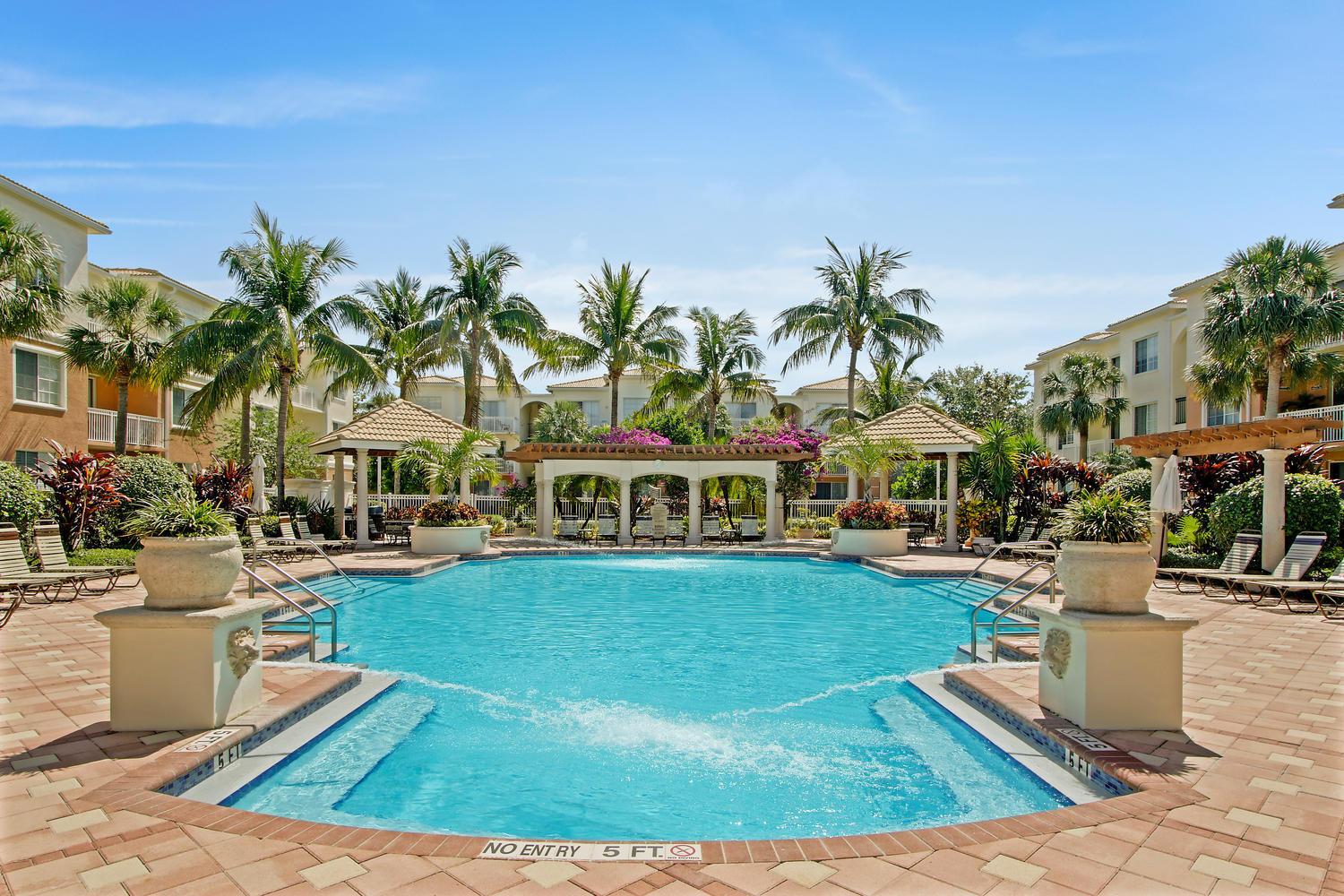 Community pool with palm trees, tiled deck, lounge chairs, and residences in the background.
