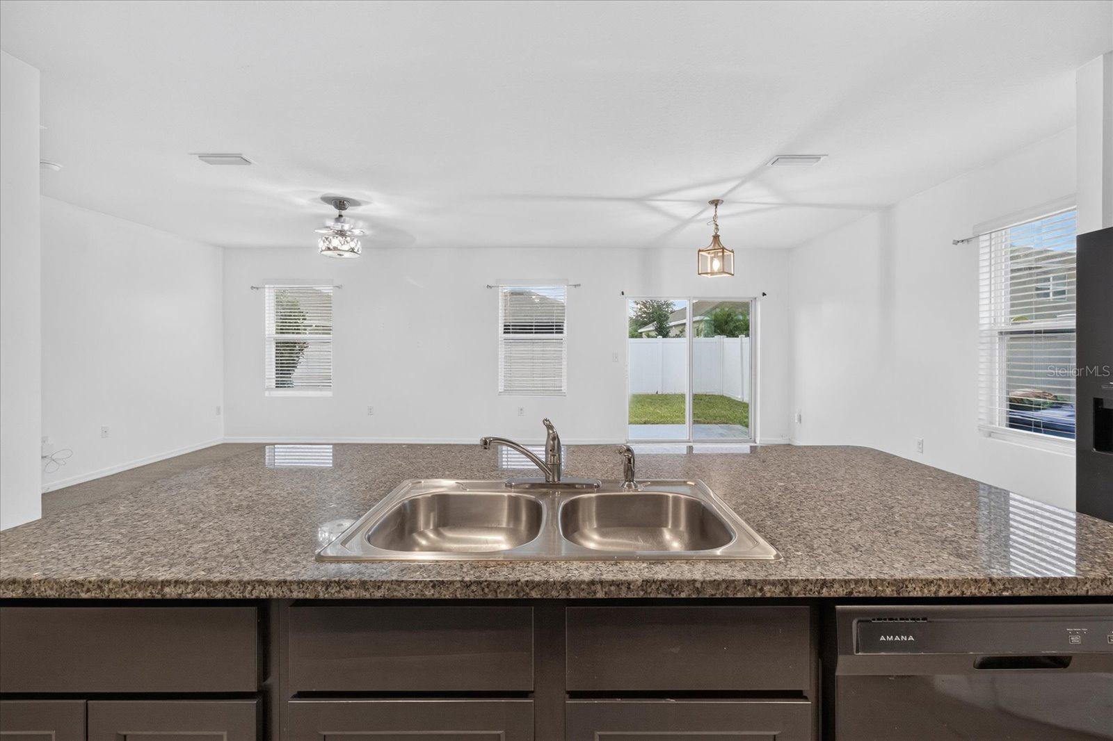 Kitchen with granite counter and double sink overlooking an unfurnished living area and fenced backyard.