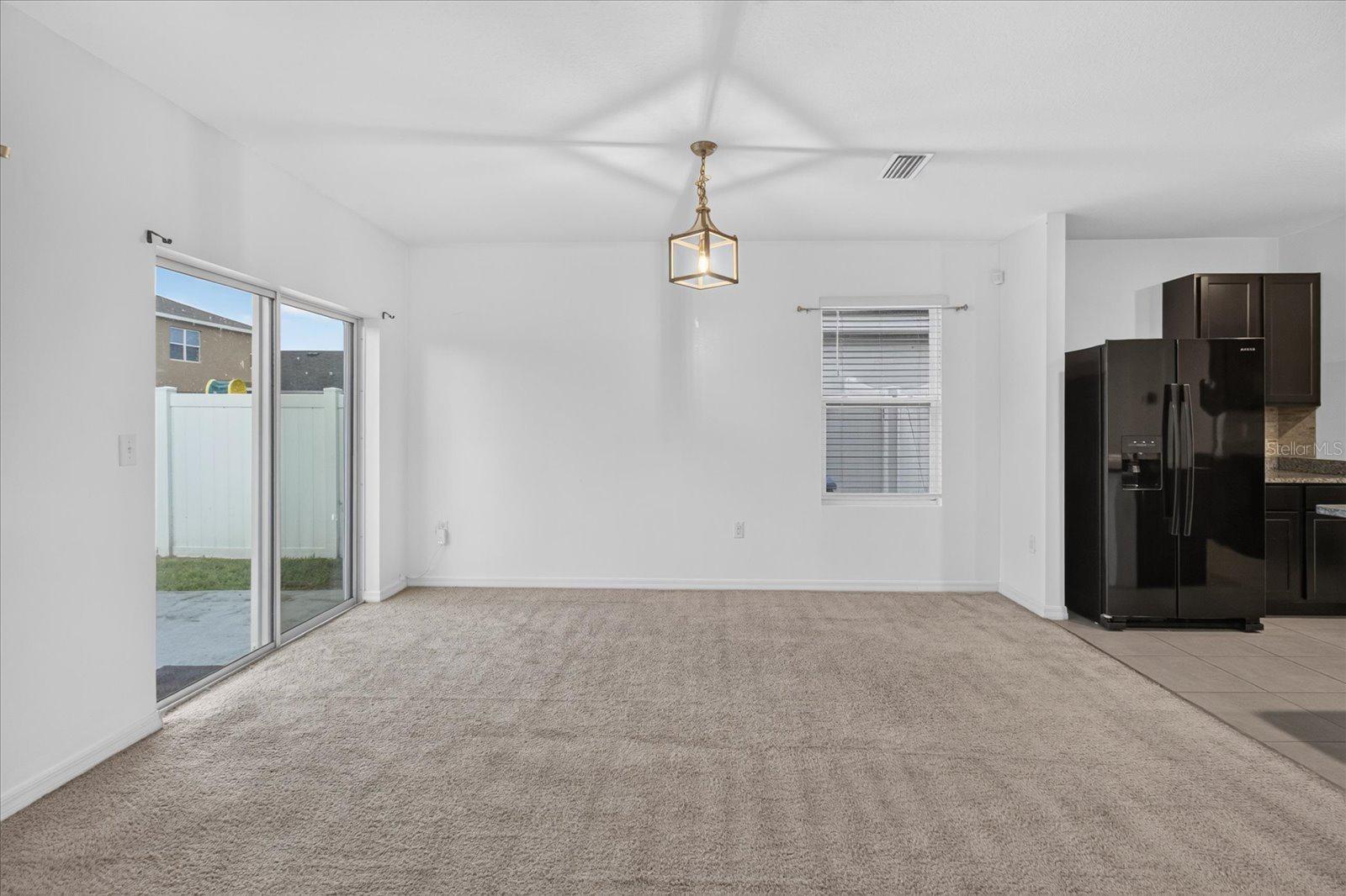 Dining area with beige carpet and sliding glass door beside a kitchen with dark cabinets and black refrigerator.