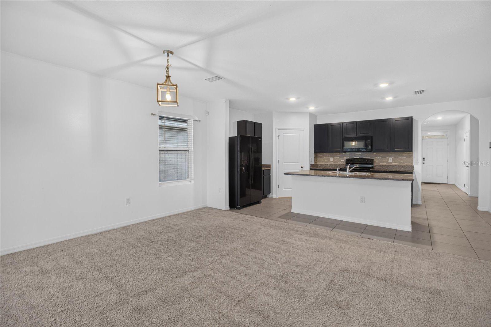 Open living area with beige carpet adjoining a kitchen with dark cabinets and a breakfast bar.